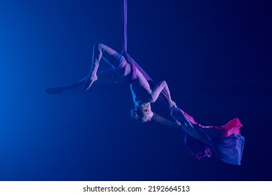 Female circus gymnast hanging upside down on aerial silk on black background with blue backlight. Young woman performs tricks at height on silk fabric. Difficult acrobatic stunts.
