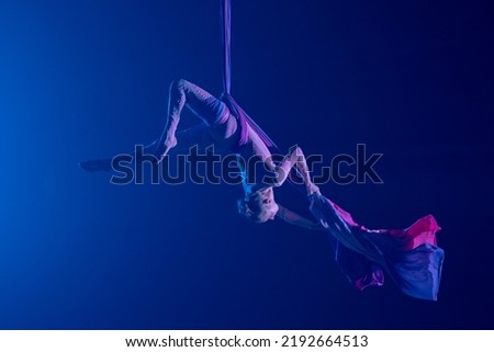 Female circus gymnast hanging upside down on aerial silk on black background with blue backlight. Young woman performs tricks at height on silk fabric. Difficult acrobatic stunts.