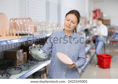 Female client near display case with kitchen utensils chooses beautiful clay plates with pattern. Customer buys view windowshopping and examines ceramic saucers and dishes