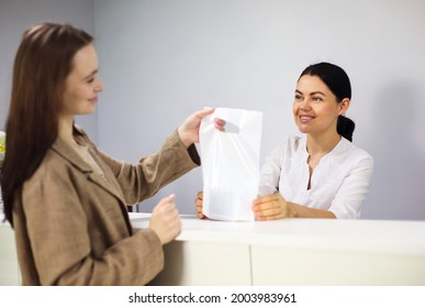 Female Client Taking Bag With Cosmetic Supplies From Friendly Employee On Reception Desk In Cosmetology Clinic