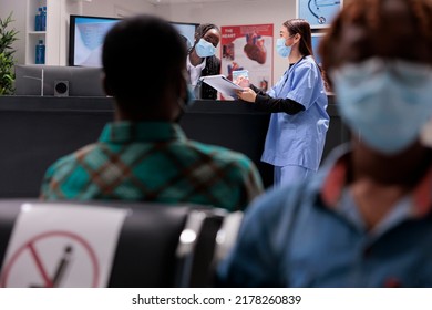 Female Clinic Receptionist Talking To Asian Nurse At Reception Desk In Hospital Lobby, Chatting About Healthcare Service And Medical Appointment. Facility Staff During Covid 19 Epidemic.