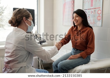 Female clinician shaking hand of young happy Hispanic patient sitting on couch in medical office after ultrasound examination