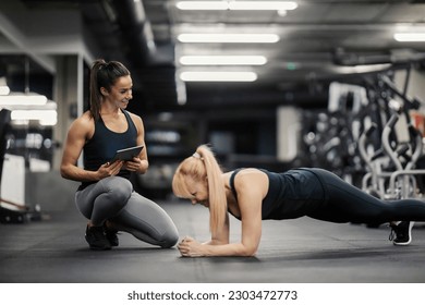 A female coach is looking at the sportswoman in a gym who is doing planks and tracking her progress on a tablet. - Powered by Shutterstock