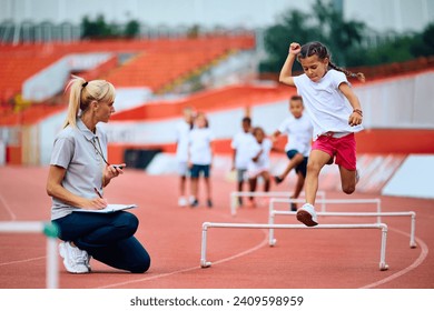 Female coach measuring time on stopwatch while girl is jumping over obstacles on running track at the stadium.  - Powered by Shutterstock