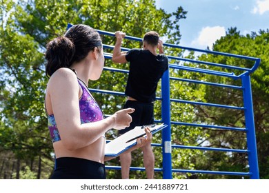 Female coach with a stopwatch and clipboard monitoring a male athlete's pull-up performance at an outdoor exercise station. - Powered by Shutterstock