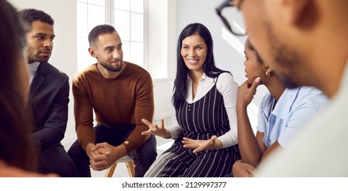 Female coach or team leader tells funny story or joke to diverse team during work meeting. Multiracial employees sitting in circle on chairs during informal brainstorming exchange ideas.