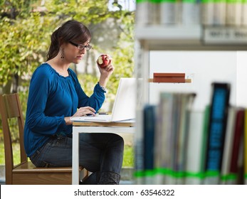 Female College Student Studying In Library And Eating Red Apple. Horizontal Shape, Side View, Three Quarter Length