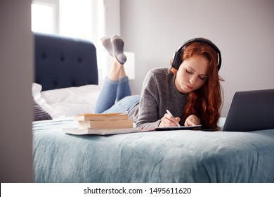 Female College Student Wearing Headphones Lies On Bed In Shared House With Mobile Phone Using Laptop