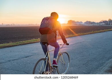 Female Commuter Riding A Bike Out Of Town To A Suburban Area. Woman Cycling Along The Road At Sunset