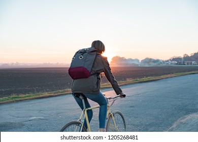 Female Commuter Riding A Bike Out Of Town To A Suburban Area. Young Woman Goes Home By Bike From Work Along The Road At Sunset