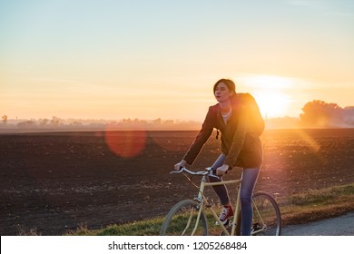 Female Commuter Riding A Bike Out Of Town. Woman Cycling Along The Road At Sunset