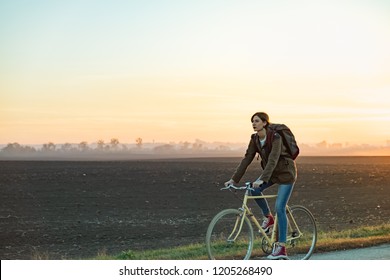 Female Commuter Riding A Bike Out Of Town In Rural Area. Young Woman Riding Bike At Sunset