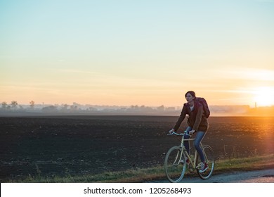 Female Commuter Riding A Bike Out Of Town In Rural Area. Young Woman Riding Bike At Sunset