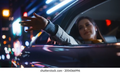 Female Is Commuting Home In A Backseat Of A Taxi At Night. Passenger Chilling And Holding Her Hand Outside Of Window While In A Car In Urban City Street With Working Neon Signs.