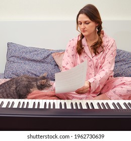 A Female Composer In Pajamas And A Cat Writes Sheet Music On Paper. Writing Music At Home With A Digital Piano