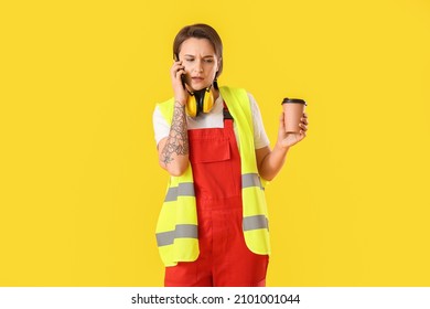 Female Construction Worker With Cup Of Coffee Talking By Mobile Phone On Yellow Background