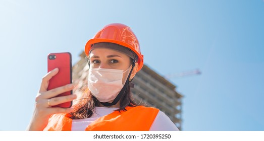 Female Construction Worker In Overalls And Medical Mask Flipping Through News About Coronavirus In Mobile Phone On Background Of House Under Construction. Concept Of Threat Of Infection.