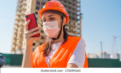 Female Construction Worker In Overalls And Medical Mask Flipping Through News About Coronavirus In Mobile Phone On Background Of House Under Construction. Concept Of Threat Of Infection.