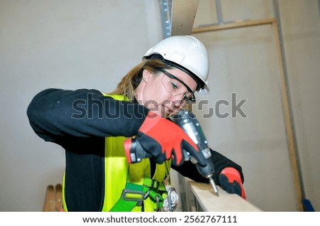 Female construction worker using power drill, mounting plasterboard on a construction site, wearing safety helmet, glasses and gloves