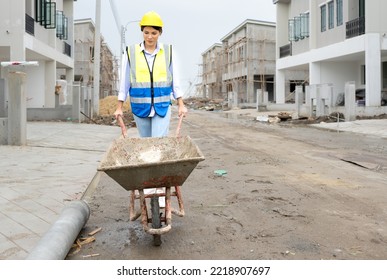 Female Construction Worker Is Working At Construction Site Pushing Wheelbarrow. Young Asian Foreman Woman In Reflective Clothing Safety Works Hard At Residential Building Development With Pushcart.