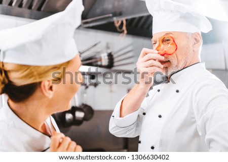 female cook looking at male chef holding pepper slice in front of face in restaurant kitchen