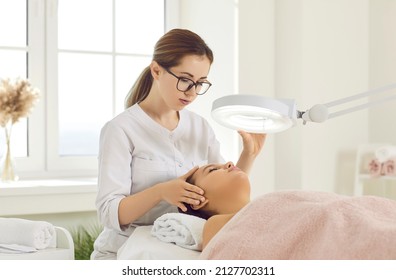 Female cosmetologist performs facial examination procedure at cosmetology clinic. Beautician fixes lamp over face of female patient lying on examination couch in beauty clinic. Cosmetology concept.