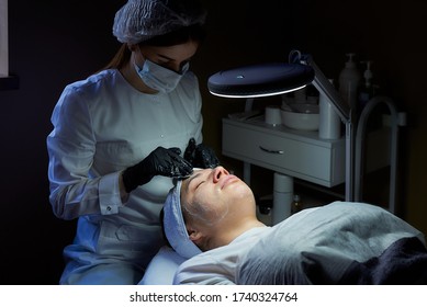 A female cosmetologist wearing a surgical face mask and disposable medical gloves rubs cream on a woman's face to tone her skin. A cosmetology procedure in a beauty salon for skin cleaning.