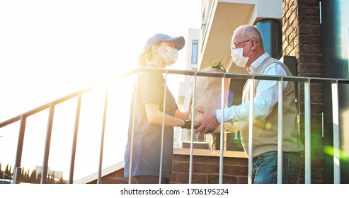 Female courier in medical mask and gloves bringing packet with fresh food to retired old man. Young Caucasian woman handing bag with grocery to male pensioner outdoor at house. Delivery service
