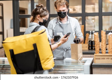 Female Courier Taking Online Order For Delivery At The Counter With Salesperson In The Small Pastry Shop Or Cafe. Concept Of A Take Away Food During Pandemic