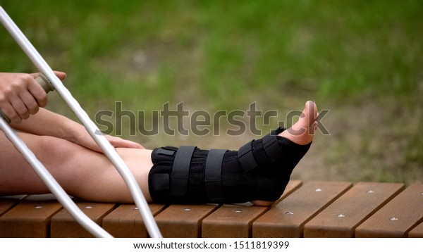 Female with crutch sitting bench with ankle brace
on leg, bone fracture,
strain