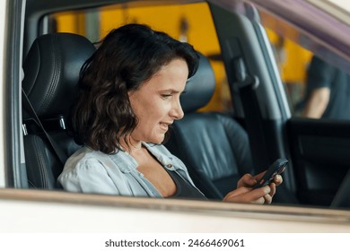 Female customer in blue denim shirt sitting in car, smiling and looking at smartphone. Background includes garage setting. Represents communication or transaction related to car repair services. - Powered by Shutterstock