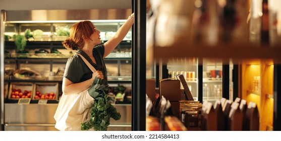 Female customer choosing food products from a shelf while carrying a bag with vegetables in a grocery store. Young woman doing some grocery shopping in a trendy supermarket. - Shutterstock ID 2214584413