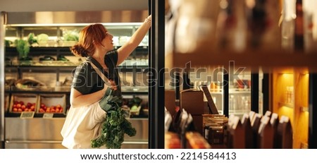 Female customer choosing food products from a shelf while carrying a bag with vegetables in a grocery store. Young woman doing some grocery shopping in a trendy supermarket.