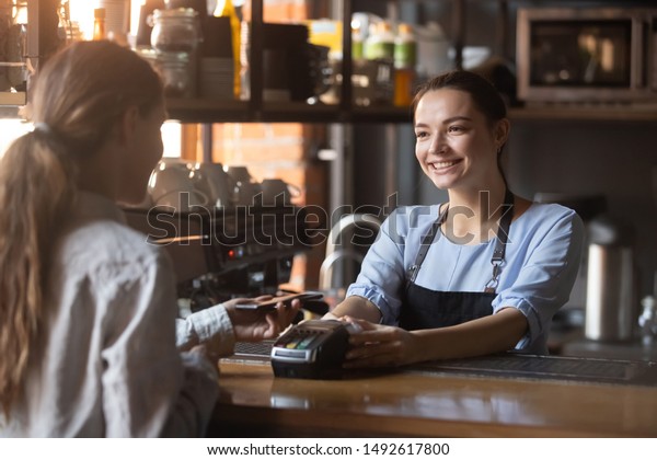 Female customer holding phone near nfc terminal\
make contactless mobile payment with smiling waitress barista\
saleswoman on coffeeshop counter, woman client pay in cafe with\
cellphone via pos\
machine