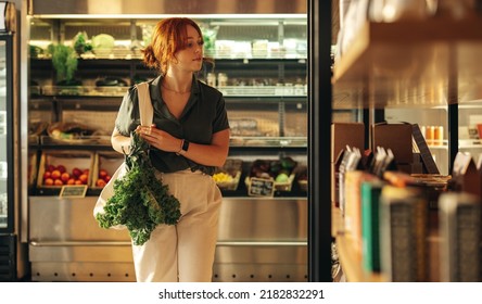 Female Customer Looking Through A Food Shelf While Carrying A Bag With Fresh Organic Vegetables In A Grocery Store. Young Woman Doing Some Grocery Shopping In A Trendy Supermarket.