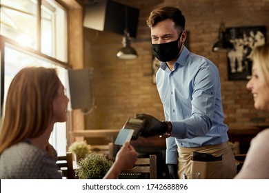 Female customer making contactless payment to a waiter who is wearing protective face mask in a cafe. 