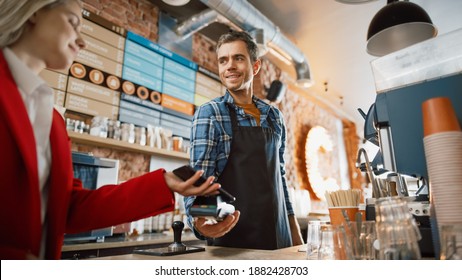 Female Customer Pays for Take Away Coffee with Contactless NFC Payment Technology on Smartphone to a Handsome Barista in Checkered Shirt in Cafe. Customer Uses Mobile to Pay Through Bank Terminal. - Powered by Shutterstock