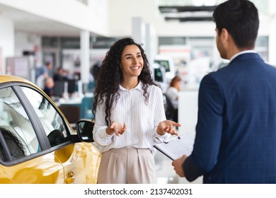 Female Customer Pretty Arab Lady With Long Curly Hair Having Conversation With Sales Associate Unrecognizable Man In Suit At Auto Dealership, Young Woman Buying Car At Modern Showroom, Copy Space