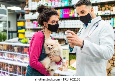 Female Customer With Protective Face Mask Talking With Veterinarian In Pet Shop And Holding Cute Pomeranian Dog.