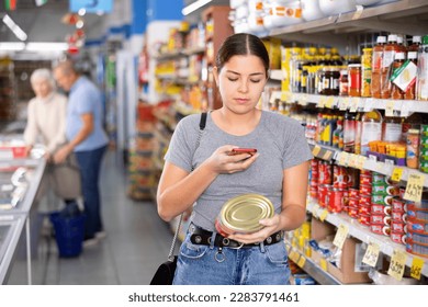 Female customer scanning barcode on aluminum canned food with smartphone while shopping in supermarket, paying for item using mobile app