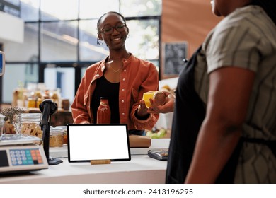 Female customer shopping at eco-friendly grocery store, for fresh organic food. Digital tablet showing blank chromakey mockup template is horizontally placed on counter while cashier assists client. - Powered by Shutterstock