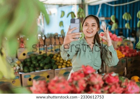 female customer standing with oke hand gesture while taking selfie photo at the fruit shop using the phone