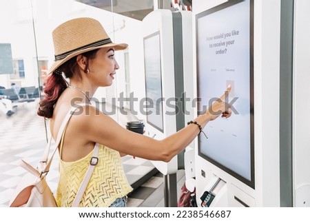 A female customer uses a touchscreen terminal or self-service kiosk to order at a fast food restaurant. Automated machine and electronic payment