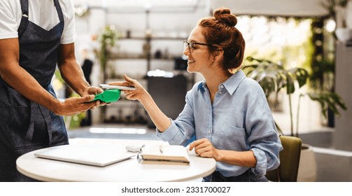 Female customer using NFC technology to pay her bill, scanning her smartphone on a payment terminal to complete the transaction. Happy woman using a cashless and contactless payment method in a cafe.