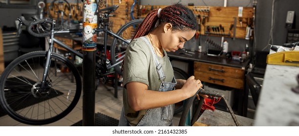 Female Cycling Master Fixing Bike Tube In Modern Bicycle Workshop. Young Concentrated Black Girl. Bike Service, Repair And Upgrade. Garage Interior With Tools And Equipment. Daytime