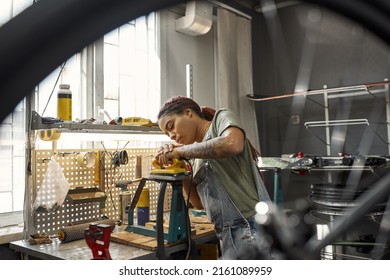 Female Cycling Repairman Fixing Bike Tube In Bicycle Workshop. Young Focused African American Girl With Tattoos. Bike Service, Repair And Upgrade. Garage Interior With Tools And Equipment