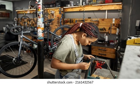 Female Cycling Repairman Fixing Bike Tube In Modern Bicycle Workshop. Young Focused African American Woman. Bike Service, Repair And Upgrade. Garage Interior With Tools And Equipment