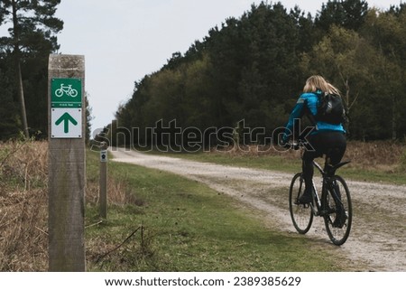 A female cyclist with blonde hair rides along a track by a cycle lane in the forest in Suffolk in England UK