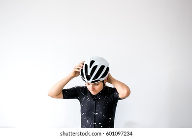 Female Cyclist Dressed In Black Standing In Front Of White Wall While Putting On Helmet