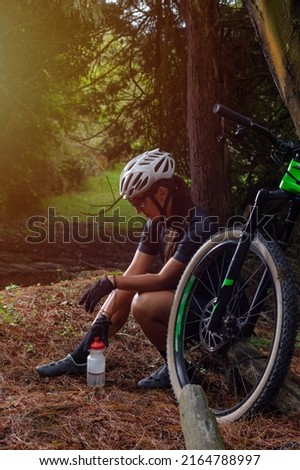 Female cyclist in the middle of the mountain sitting on a log holding a bottle of water while her bike is leaning against a tree